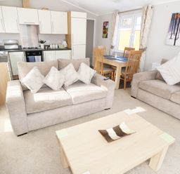 A living area with beige sofas a wooden coffee table a dining table with chairs and a kitchen with white cabinets at 32 Cruachan Lodge in Auchterarder