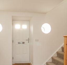 An entryway with a white front door two round windows a wooden side table with a lamp and stairs with a wooden banister at Coney Garth Barn in Beckermet