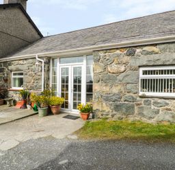 Stone exterior of a house with white framed glass double doors and potted plants on the paved area at Bwthyn Ael Y Bryn in Criccieth