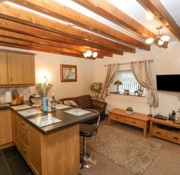 A kitchen and living area with wooden cabinets a countertop with stools a brown sofa a rug and a wall-mounted TV at Bwthyn Ael Y Bryn in Criccieth
