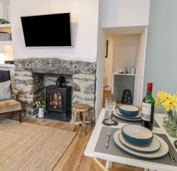 A living room with a stone fireplace stove a mounted TV an armchair and a dining table set with dishes wine and flowers at Llys Madoc in Porthmadog