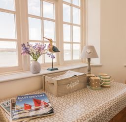 A dining table with cups, magazines, a wooden crate, a lamp, flowers, and bird figurines by large windows at Searles in Lympstone