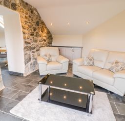 A living room with a leather sofa and armchair a black glass coffee table and a stone wall at Bothi in Llanuwchllyn