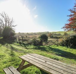 A wooden picnic table on grass near a dirt path with a wooden gate and fields in the background at Pengraig near Tregaron