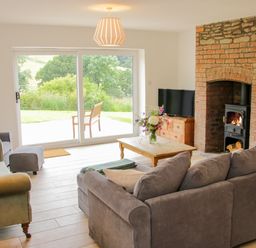 A living room with a gray sofa green armchairs a wooden coffee table flowers a brick fireplace and sliding glass doors showing a garden at The Old Farmhouse Upper Pitts in Knighton