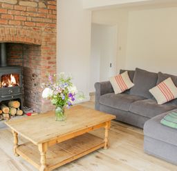 A living room with a wooden coffee table with flowers a gray sofa with cushions and a wood burning stove in a brick fireplace at The Old Farmhouse Upper Pitts in Knighton