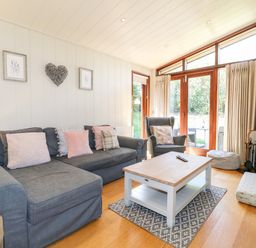 A living room with a gray sofa, armchair, wooden coffee table, rug, wood stove, and large windows with curtains at Brooke in Exton Rutland