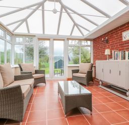 A conservatory with wicker chairs and a sofa around a glass table and a cabinet with books at Bower View in Madley