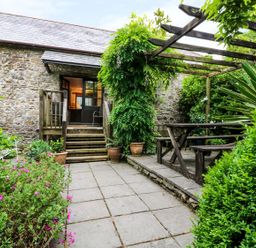 A stone patio with wooden steps leading to a door and a pergola with a picnic table surrounded by plants at The Cider Barn in Loddiswell