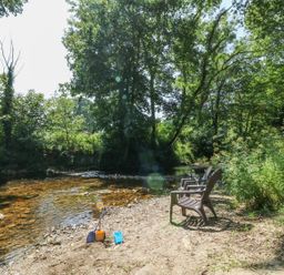 A riverbank with two chairs and children's sand toys near a flowing river surrounded by trees at The Cider Barn in Loddiswell