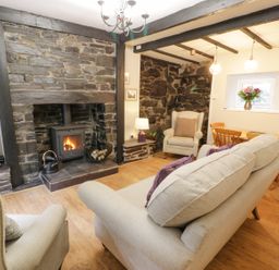 A living room with a stone fireplace a stove beige sofas a wooden dining table and chairs and flowers in a vase at Ty Coed in Llanberis