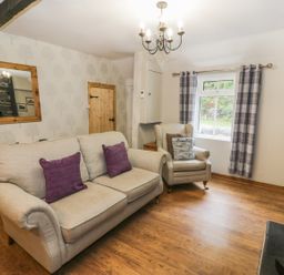 A living room with beige sofa and armchair with cushions wooden floor window with plaid curtains and a wooden door at Ty Coed in Llanberis
