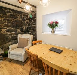 A dining area with a wooden table and chairs a beige armchair next to a stone wall and a window with flowers on the sill at Ty Coed in Llanberis