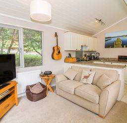 A living room with a beige sofa a wooden TV stand with a television a small wooden side table a guitar hanging on the wall and a kitchen area with white cabinets and appliances at Willow Lodge in Troutbeck Bridge