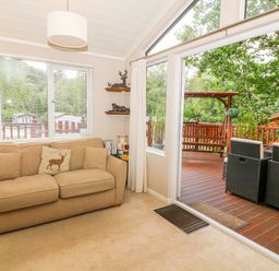 A living room with a sofa and deer cushion next to a window and glass door leading to a wooden deck with outdoor furniture at Willow Lodge in Troutbeck Bridge