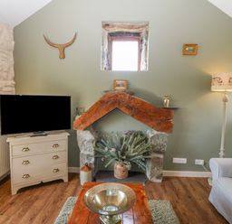 A living room with a television on a dresser stone fireplace with wood beam and plants a floor lamp and a sofa at Beudy in Chwilog