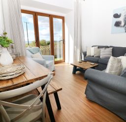 A living room with a wooden dining table with chairs a vase with flowers a blue armchair a blue sofa with cushions a small wooden coffee table and a painting of a cow on the wall at Sgubor in Chwilog
