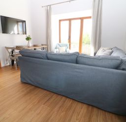 A living room with a gray sofa a wooden dining table with chairs a wall mounted television and glass doors at Sgubor in Chwilog