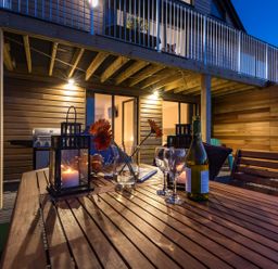 A wooden outdoor dining table with a wine bottle glasses flowers and lanterns on a patio under a balcony at Godrevy in St Ives