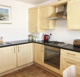 A kitchen with wooden cabinets black countertops a stainless steel sink gas stove oven toaster kettle and a framed painting at 5 Chandlers Yard in Burry Port