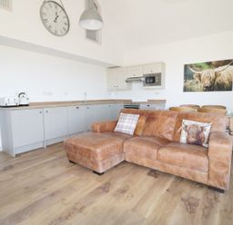 An open plan kitchen and living area with a brown leather sofa wooden dining table and chairs wall clock and kitchen appliances at Heron Lodge Castle Farm in Wedmore