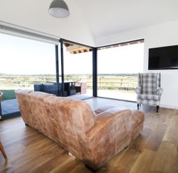 A living room with a brown couch two plaid armchairs a wall mounted TV large windows overlooking an outdoor patio at Heron Lodge Castle Farm in Wedmore