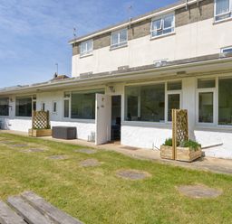 A garden area with a grass lawn and stepping stones in front of a white building with windows and an open door at 2 Brook Cottages in Hope Cove