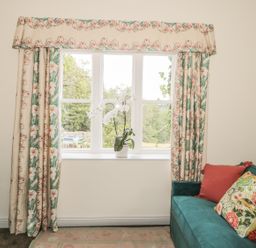 A window with floral curtains and a white orchid on the sill next to a teal sofa with colorful cushions at Newbiggin Cottage in Blanchland