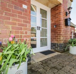The entrance of a brick building with white double doors two flower pots with pink and purple flowers on a paved ground at Carpenters Cottage in Cockermouth