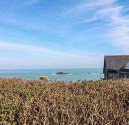 A view of the ocean with rocks and a hedge in the foreground and a house with solar panels on the roof