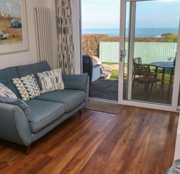 A living room with a blue sofa and geometric patterned cushions next to sliding glass doors opening to a garden and sea view at Kittiwake in Crantock