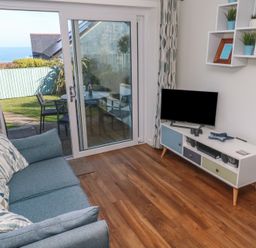 A living room with a blue sofa a TV on a white stand and sliding glass doors leading to a garden at Kittiwake in Crantock