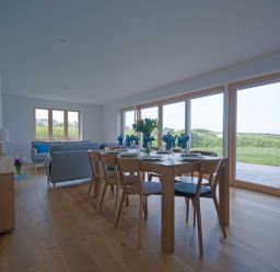 A dining table set with blue napkins and plates beside sofas and large windows showing greenery at Kareena in Crantock