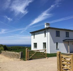 A white two-story house with multiple windows and wooden gates on a gravel driveway with a stone wall and greenery nearby and a blue sky in the background at Kareena in Crantock