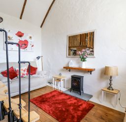 A living room with a spiral staircase a beige couch with red pillows a red rug a small black stove and a wall mirror at Rose Cottage in Penrhiwllan