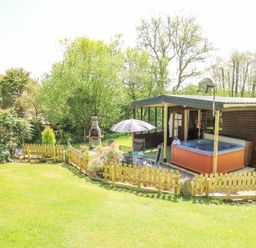 A wooden cabin with a hot tub patio umbrella chairs a small fence and a barbecue in a garden at Brook Lodge Penrhiwllan