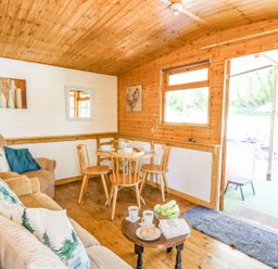 A wooden cabin living room with a sofa, armchair, small round dining table with four chairs, and open door to outside at Brook Lodge Penrhiwllan