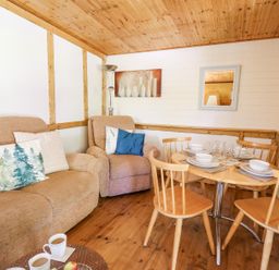 A living area with a beige sofa and armchair and a wooden dining table set with dishes and glasses at Brook Lodge in Penrhiwllan