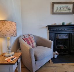 A corner with a beige armchair next to a fireplace and a side table with a lamp and a book at Moelwyn View Cottage in Llan Ffestiniog