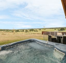An outdoor hot tub with bubbling water next to a patio dining set overlooking a fenced grassy field at Swan Lodge Castle Farm in Wedmore