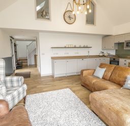 A living room with brown leather sofa, plaid armchair, white rug, and an open kitchen with wooden countertops at Swan Lodge Castle Farm in Wedmore