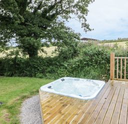 An outdoor hot tub on a wooden deck next to a grassy area with trees and bushes at Oak Lodge in Constantine near Falmouth