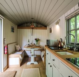 A kitchen and dining area with wooden countertops a sink induction cooktop a dining table with wine glasses and a bottle and built in benches at Rock View Shepherd's Hut in Gulworthy near Tavistock