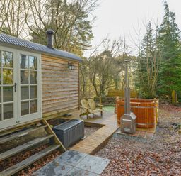 A wooden hut with glass double doors next to a deck with chairs and a wooden hot tub at Rock View Shepherd's Hut in Gulworthy near Tavistock