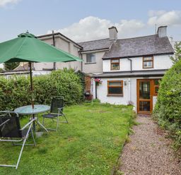 A backyard garden with a green umbrella and black chairs around a glass table next to a path leading to a white house with wooden framed windows at Gwenallt in Malltraeth