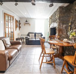 A living room with leather sofas a wooden dining table and chairs a stone wall and a glass door at Gwenallt in Malltraeth
