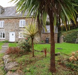 A stone house with white windows and door surrounded by a garden with palm trees and green grass at Lanvean Farmhouse Lanvean near St Mawgan