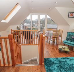 An attic living space with wooden railing stairs a dining table with four chairs a teal armchair and ottoman a blue rug and large windows at River Cottage in Morpeth