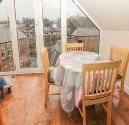 A dining area with a round table covered by a tablecloth and four wooden chairs near a glass door overlooking buildings at River Cottage in Morpeth