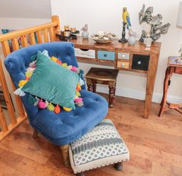 A blue chair with a colorful pillow and footstool next to a wooden table with drawers and decorative items at River Cottage in Morpeth